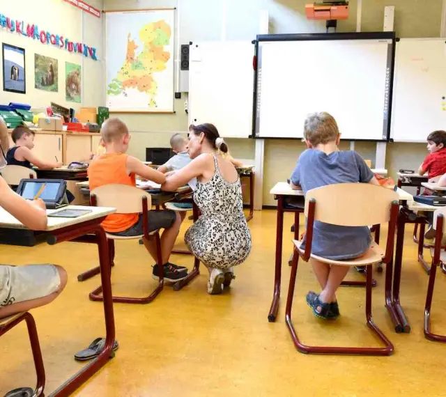 Dzieci siedzą w klasie, ucząc się. Nauczycielka pochyla się nad jednym z uczniów. Ile jest klas w Anglii? Tego nie wiemy, ale ta lekcja wygląda na interesującą.