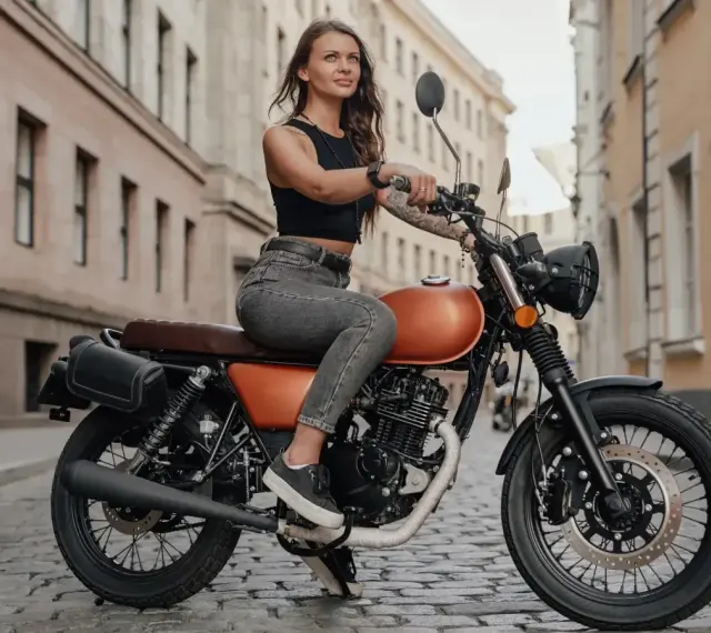 Kobieta w czarnym topie i jeansach siedzi na pomarańczowym motocyklu na brukowanej ulicy. To idealna kategoria a dla miłośniczek wolności.