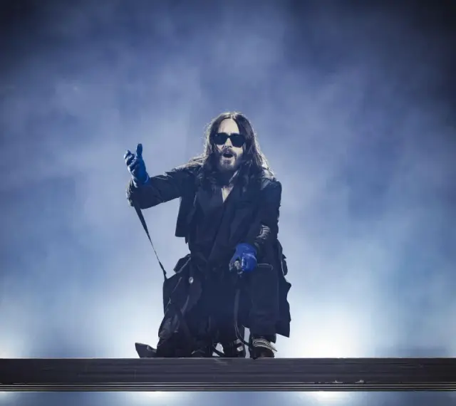 Jared Leto w niebieskich rękawiczkach, w okularach i długim płaszczu, klęczy na scenie. Jego zespół pewnie czeka na jego znak.