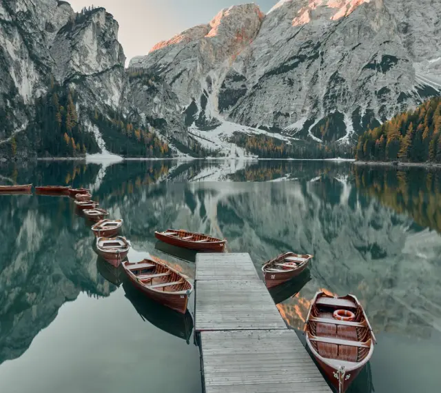 Drewniane łodzie przy drewnianym pomoście na spokojnym Lago di Braies, odbijające się w tafli wody, z majestatycznymi górami w tle.