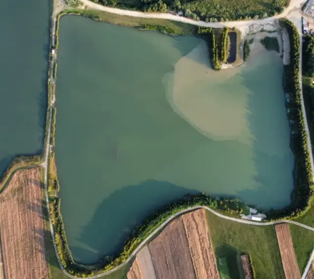 Widok z lotu ptaka na zielone oczko wodne, otoczone polami i drogami. Idealne miejsce na spokojne łowisko.