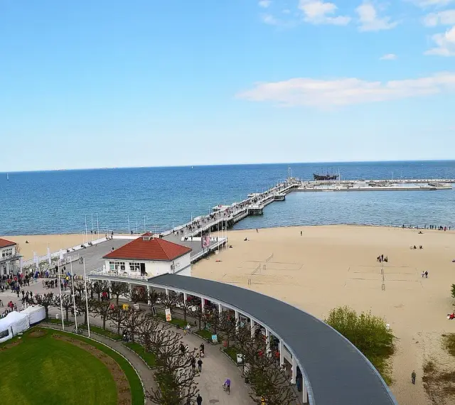 Sopot beach z molo, kawiarniami i spacerującymi ludźmi. W oddali widać statki.