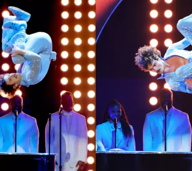 Wokalista w błyszczącym niebieskim stroju wykonuje akrobacje na scenie podczas gali nagród Grammy. Laureaci nagród grammy w białych garniturach stoją za fortepianami.