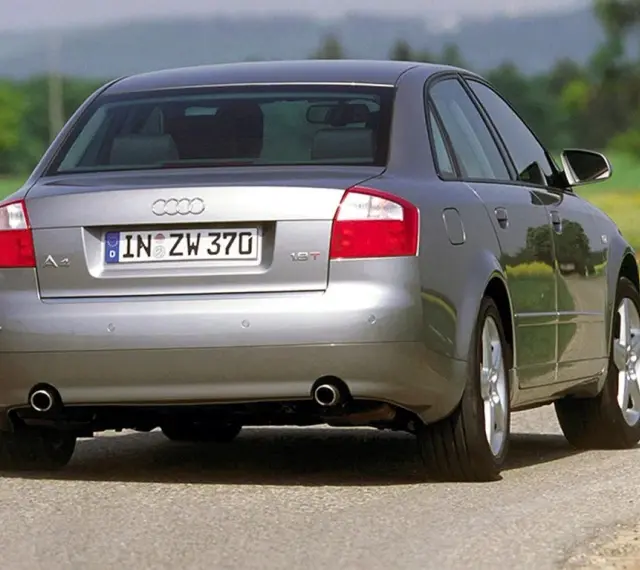 Srebrne Audi B6 sedan z tablicą rejestracyjną IN ZW 370 jedzie po asfaltowej drodze wśród zieleni i żółtych kwiatów.