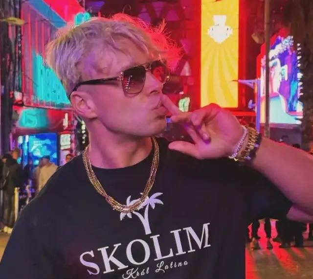 Skolim w okularach i złotych łańcuchach, pozuje na tle neonów. Król Latino w swoim wieku, w blasku miasta.
