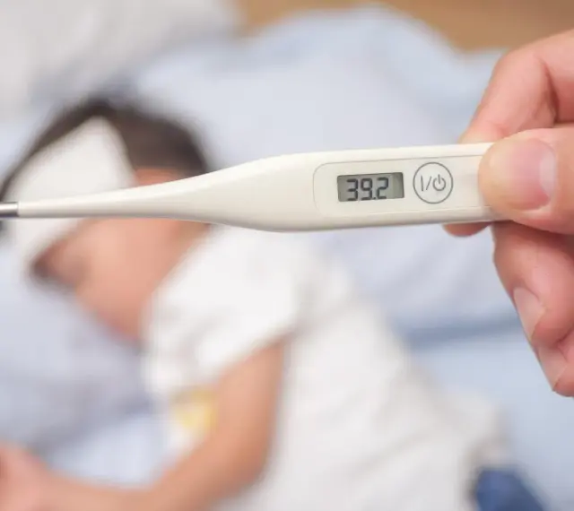 Cyfrowy termometr pokazuje 39.2°C, wskazując wysoką temperaturę ciała u chorego dziecka leżącego w łóżku.