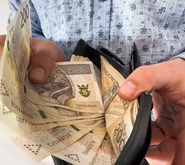 Dłonie trzymają portfel z banknotami 200 zł. Ile trzeba przepracować, by zasłużyć na taką 13 pensję?