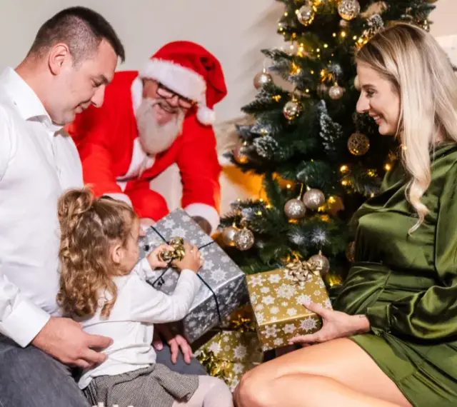 Rodzina otwiera prezenty przy choince, a Mikołaj uśmiecha się w tle. Wesołe święta w hotelu.