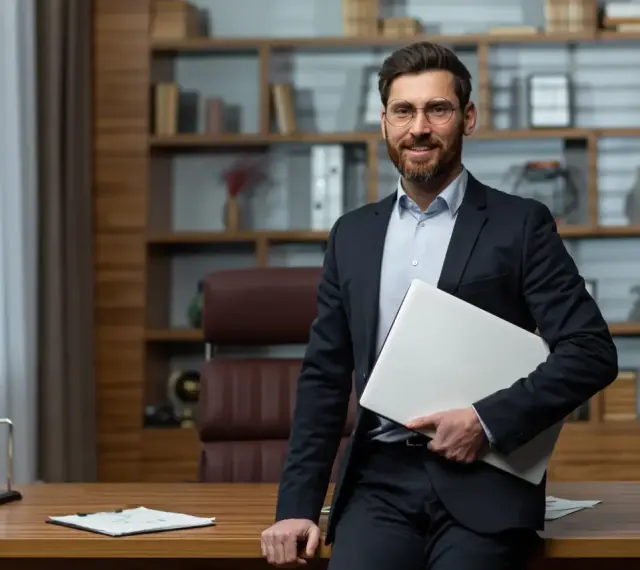 Uśmiechnięty dyrektor szkoły w garniturze, trzymający laptopa, stoi przy biurku w gabinecie.