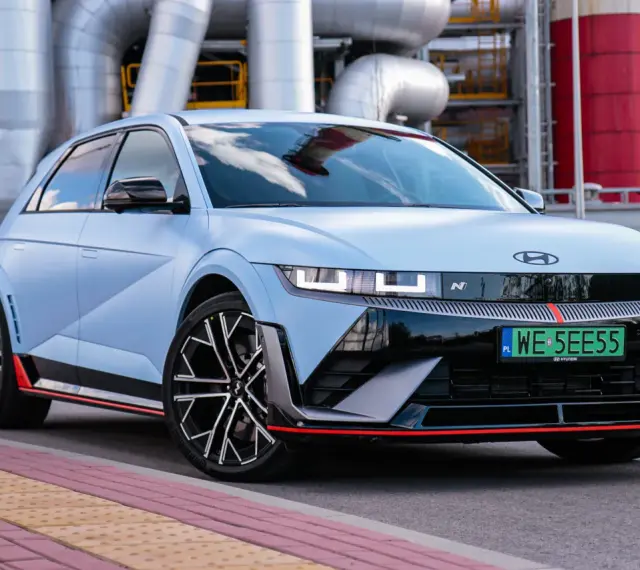 Jasnoniebieski Hyundai IONIQ 5 N z czarnymi akcentami i czerwonymi detalami, zaparkowany przed przemysłową instalacją.