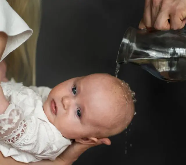 Chrzest dziecka. Woda spływa po głowie malucha w białej szatce. Niech to będzie początek mądrych życzeń na chrzest.