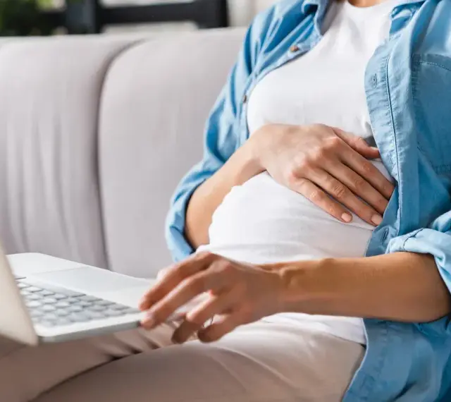 Kobieta w ciąży siedzi na kanapie, pisząc na laptopie wniosek o urlop macierzyński.