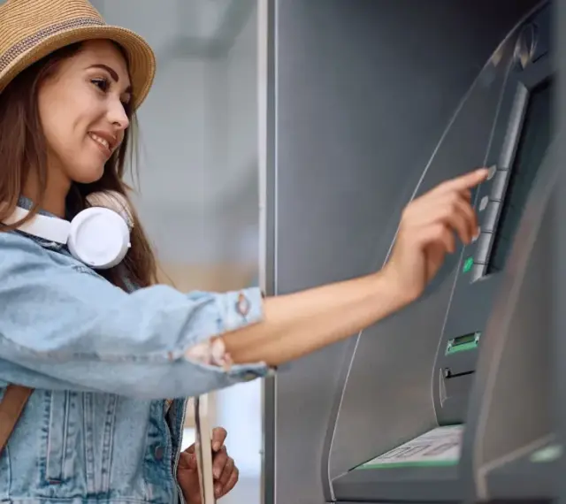 Kobieta w słomkowym kapeluszu i dżinsowej kurtce korzysta z bankomatu.