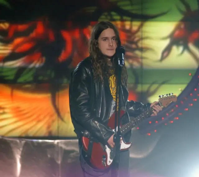Zalewski w Idolu, z gitarą i w skórzanej kurtce, na tle kolorowej scenografii.