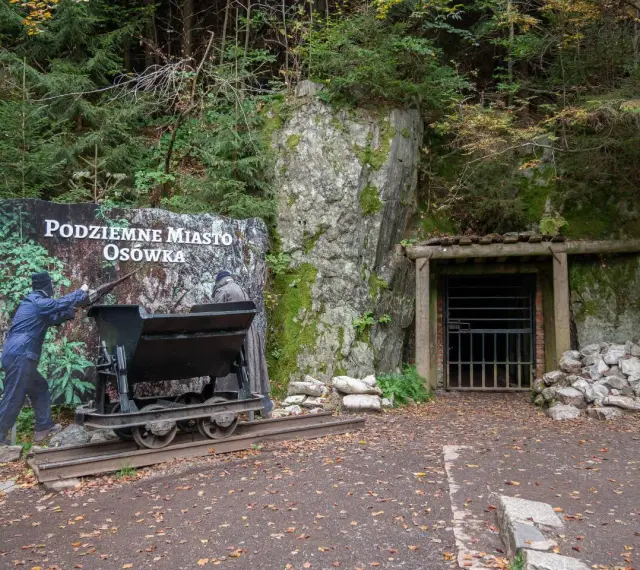 Wejście do Podziemnego Miasta Osówka, z wagonikiem i tablicą informacyjną. Widać skalne ściany i wejście do tunelu.