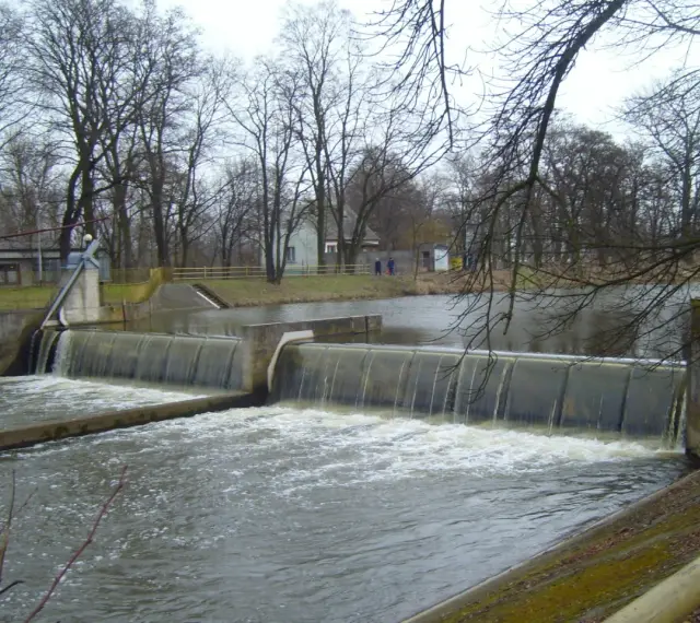 Zapora na rzece z wodospadem, drzewa bez liści, budynki w tle.
