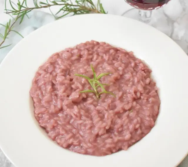 Różowe risotto z prawdziwkami i rozmarynem na białym talerzu, obok widelca i kieliszka wina.