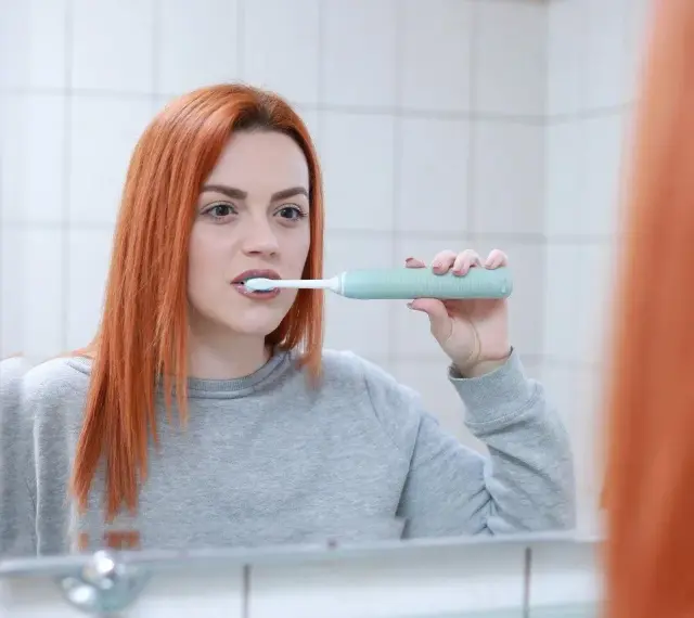 Rudowłosa kobieta myje zęby szczoteczką soniczną, zastanawiając się, czy szczoteczka soniczna usuwa kamień.