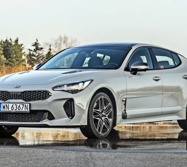 Elegancka Kia Stinger 2025 w kolorze srebrnym, zaparkowana na mokrej nawierzchni, z drzewami w tle.