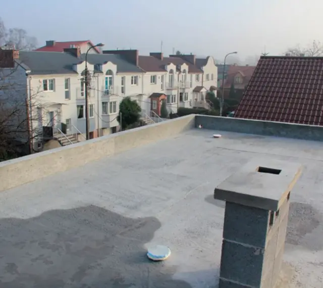 Nowy, betonowy dach płaski z kominem i widokiem na szereg domów z dachówką.