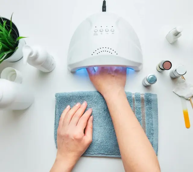 Paznokcie hybrydowe krok po kroku: dłoń w lampie UV, obok kosmetyki i akcesoria do manicure.
