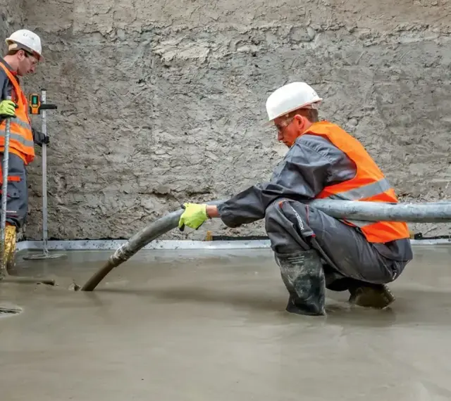 Dwóch budowlańców w kamizelkach odblaskowych wylewa pianobeton. Jeden kieruje wężem strumień materiału, drugi kontroluje poziom.