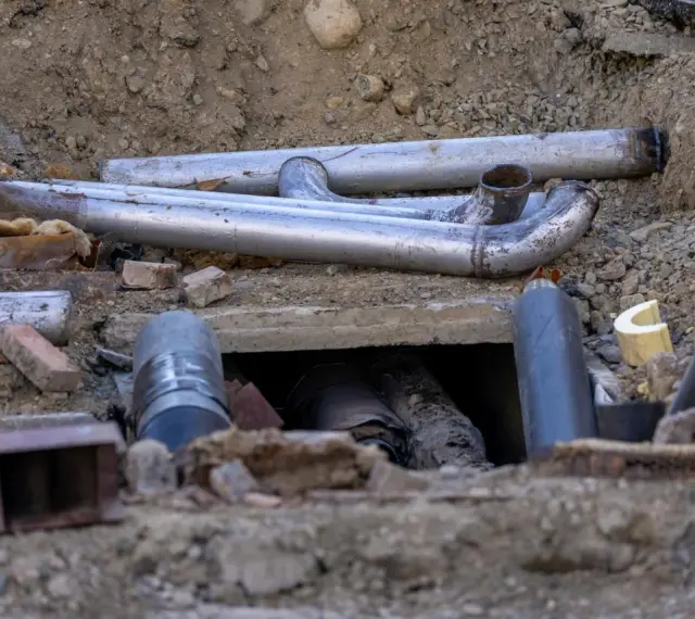 Metalowe rury tworzą skomplikowane przyłącze kanalizacyjne w wykopie. Widać fragmenty izolacji i gruzu.