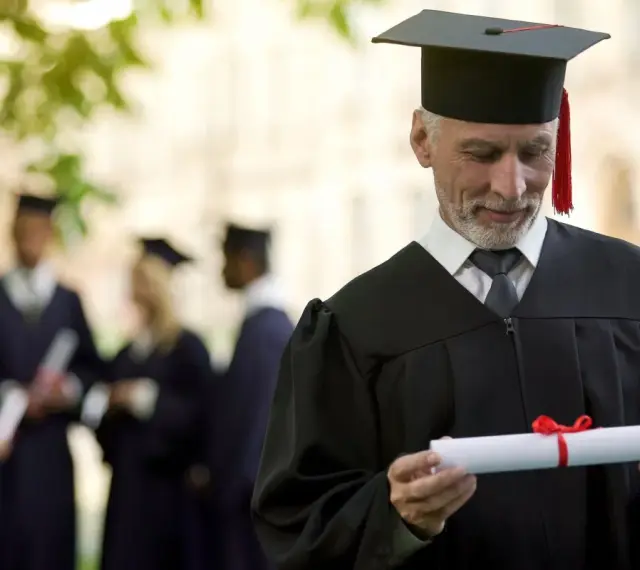 Starszy pan w todze z dyplomem zastanawia się, czy studia liczą się do stażu pracy. W tle inni absolwenci.