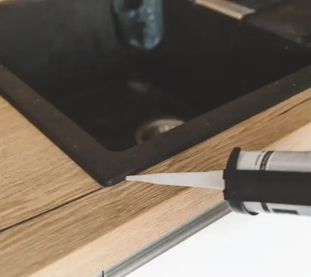Pistolet z silikonem aplikuje uszczelnienie wokół zlewu, pokazując, jak polożyć silikon na blacie kuchennym.