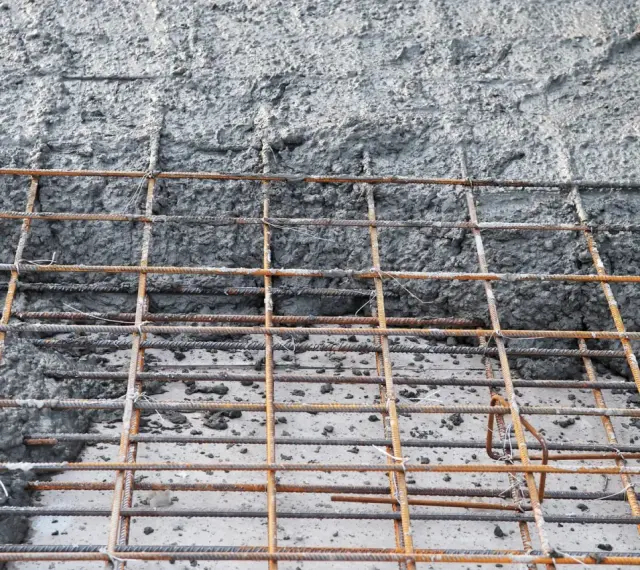 Siatka zbrojeniowa czeka na beton na fundament. Powierzchnia jest szara i chropowata, z widocznymi prętami.