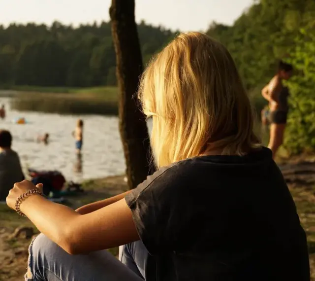 Dziewczyna z blond włosami siedzi nad jeziorem Trzechowskim, obserwując ludzi kąpiących się w wodzie.