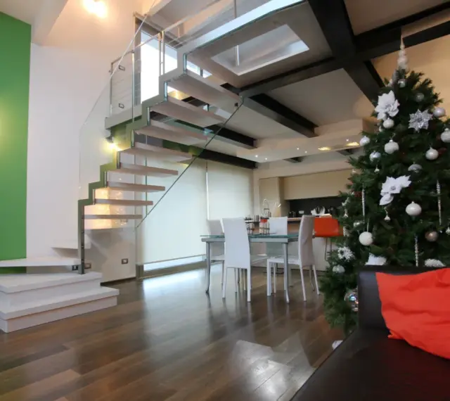 Un moderno soppalco casa con scala in vetro e legno, albero di Natale addobbato e un divano con cuscino arancione.