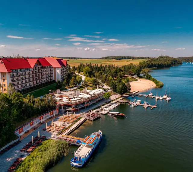 Hotel na wyspie Mikołajki z czerwonym dachem, otoczony zielenią, z mariną pełną łodzi i statkiem wycieczkowym na jeziorze.