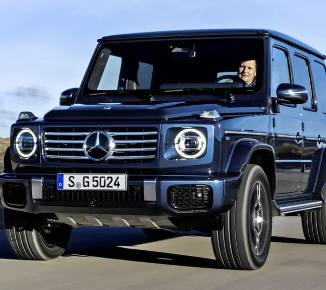 Ciemnoniebieski Mercedes G Klasa jedzie po pustynnej drodze. Kierowca uśmiecha się. Sprawdź mercedes g klasa cena!