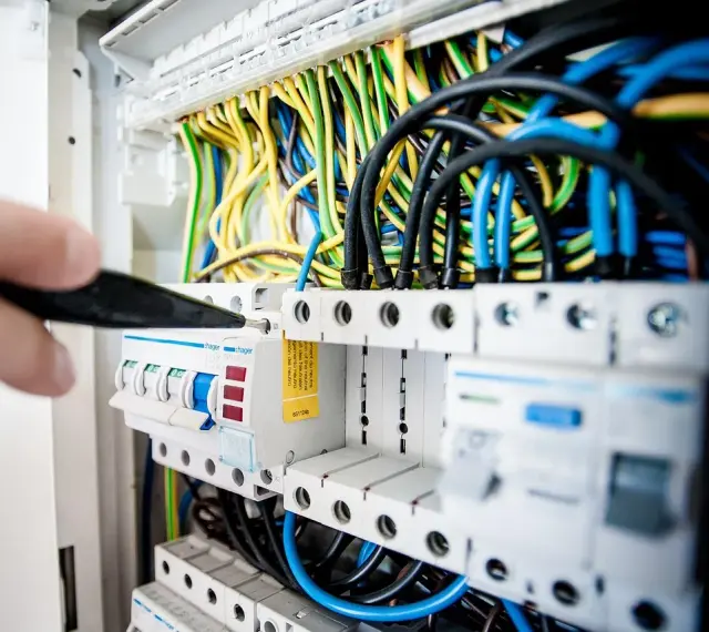 Ręka majstruje przy bezpiecznikach. Kolorowe przewody tworzą skomplikowaną instalację elektryczną.