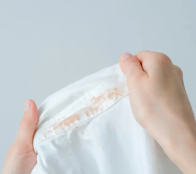 Dłonie trzymają białą koszulę z plamami po fluidzie. Jak sprać fluid? Podpowiedź na zdjęciu.