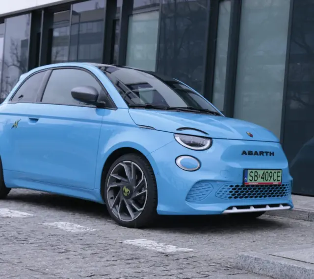 Niebieski Abarth 500e, gotowy do testów, zaparkowany przed nowoczesnym budynkiem.