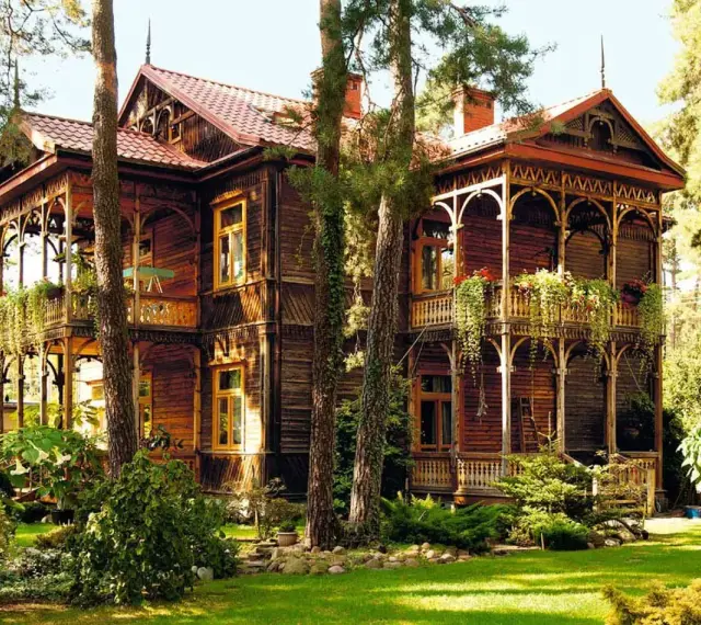 Drewniana willa w stylu świdermajer, otoczona zielenią, z pięknymi werandami i zdobieniami.