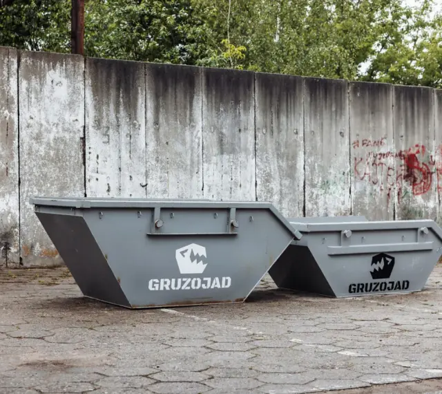 Dwa szare kontenery na śmieci zmieszane, z logo GRUZOJAD, stoją przed zniszczonym betonowym murem pokrytym graffiti.