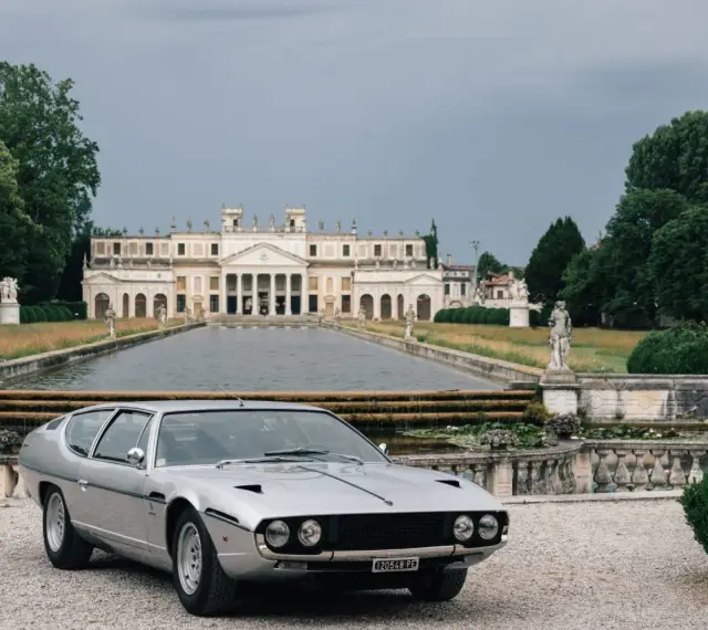 Srebrny Lamborghini Espada z lat 70. przed okazałą willą z basenem i rzeźbami.