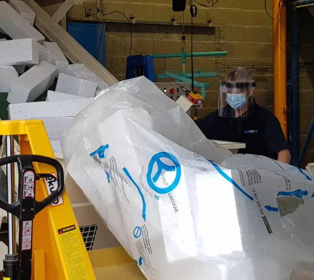Pracownik w masce i przyłbicy przygotowuje styropian do recyklingu. Duży worek z logo Knauf Circular symbolizuje proces recyklingu styropianu.