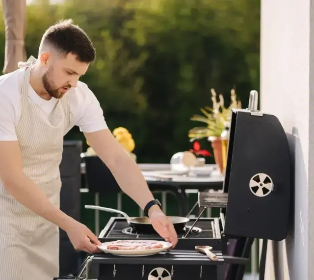 Mężczyzna w fartuchu przygotowuje mięso na grillu na balkonie. Czy można robić grilla na balkonie? Tak, jeśli jest bezpiecznie!