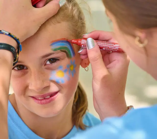 Une petite fille souriante se fait maquiller sur la joue un arc-en-ciel et des fleurs, au stand maquillage d'une kermesse.