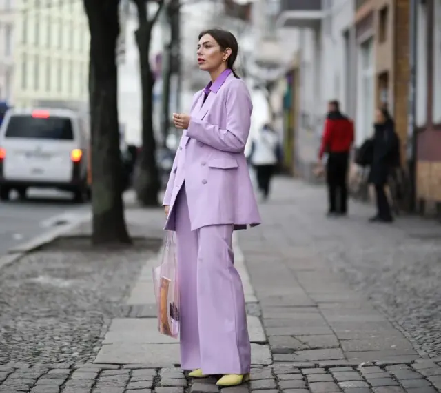 Elegancka kobieta w garniturze w kolorze liliowym, z transparentną torbą, idzie ulicą.