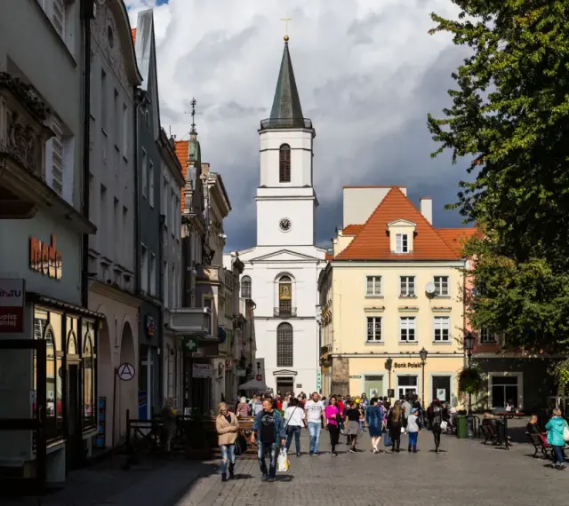 Ulica w jednym z miast na zachodzie Polski, z kościołem w tle i ludźmi spacerującymi po rynku.