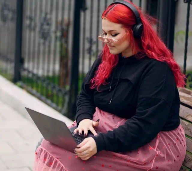 Młoda babista z ognistymi włosami i słuchawkami na uszach pracuje na laptopie, siedząc na ławce w parku.
