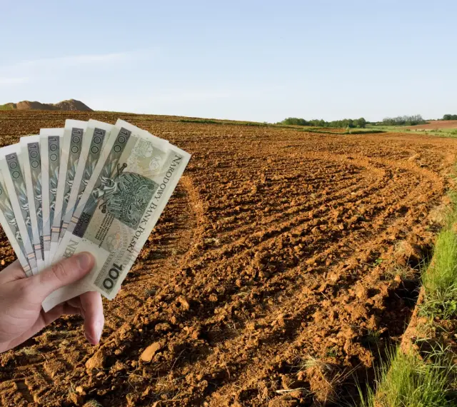 Dłoń trzyma banknoty 100 zł na tle zaoranego pola. Zastanawiasz się, ile kosztuje ar ziemi rolnej?
