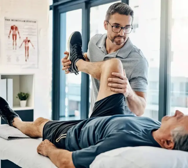 Ein Physiotherapeut dehnt das Bein eines Patienten. Der Patient liegt auf einer Liege, während der Therapeut sein Knie beugt. Wann Physiotherapie? Hier wird geholfen.