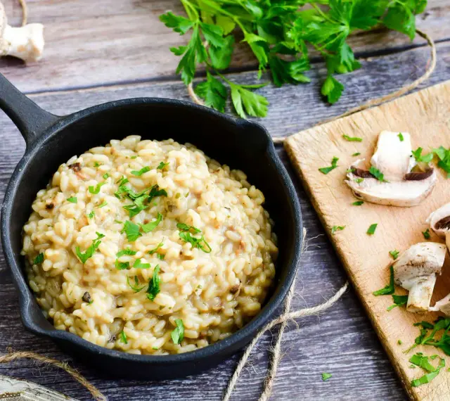 Kremowe risotto z grzybami w czarnej patelni, ozdobione natką pietruszki, obok świeże grzyby na desce.