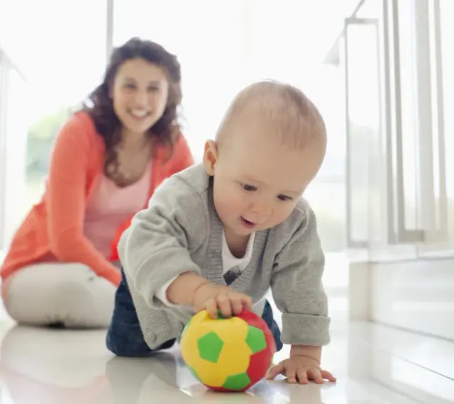 Maluch ćwiczy motorykę dużą, raczkując po podłodze i bawiąc się kolorową piłeczką. Uśmiechnięta mama obserwuje jego postępy.